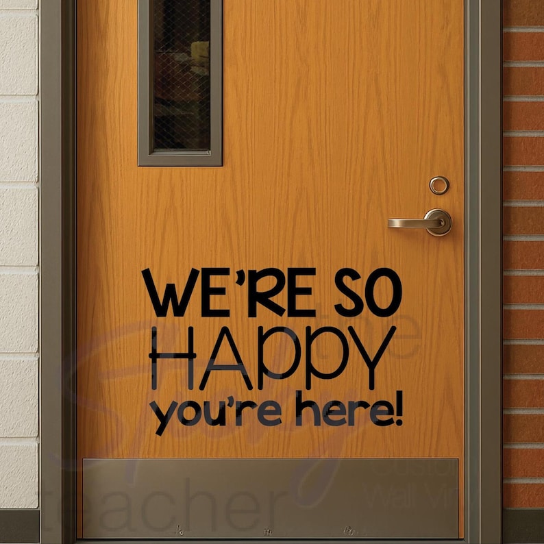 We're So Happy You're Here decal on wooden door beside brick wall